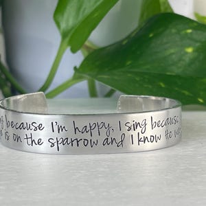 K&ouml;nnte beinhalten: Ein silberfarbenes Armreif-Armband mit eingraviertem Text. Der Text lautet: &bdquo;Ich singe, weil ich gl&uuml;cklich bin, ich singe, weil er auf dem Spatz ist und ich wei&szlig;, dass er zuschaut.&ldquo; Das Armband wird vor gr&uuml;nem Laub gezeigt.