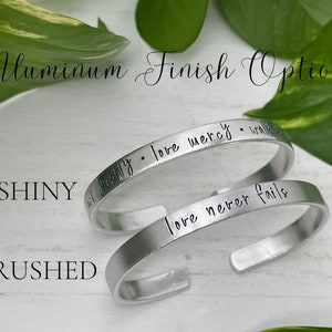May include: Two aluminum bracelets with different finishes. The top bracelet is shiny and has the text "All I truly love mercy - walk in love" engraved on it. The bottom bracelet is brushed and has the text "love never fails" engraved on it.
