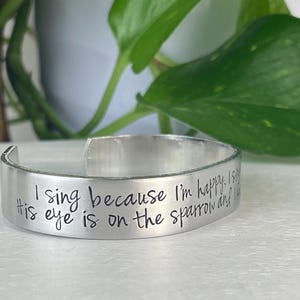 K&ouml;nnte beinhalten: Ein silbernes Armreif-Armband mit dem eingravierten Text "I sing because I'm happy. His eye is on the sparrow and I know He watches me." Das Armband ist offen und liegt auf einer wei&szlig;en Oberfl&auml;che.
