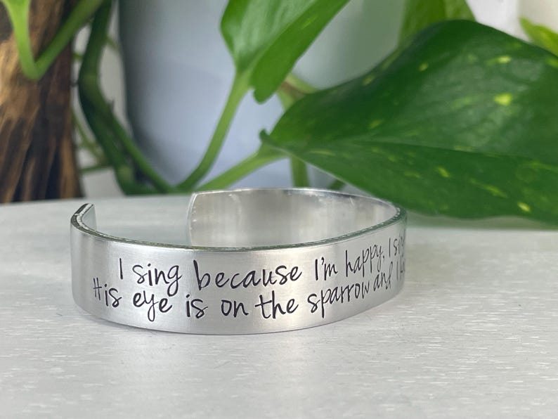 K&ouml;nnte beinhalten: Ein silbernes Armreif-Armband mit dem eingravierten Text "I sing because I'm happy. His eye is on the sparrow and I know He watches me." Das Armband ist offen und liegt auf einer wei&szlig;en Oberfl&auml;che.