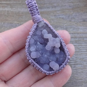 May include: A teardrop-shaped pendant featuring a cluster of small, rounded, light purple grape agate crystals. The stone is encased in a woven, light purple macrame cord frame that extends into a loop at the top for hanging.