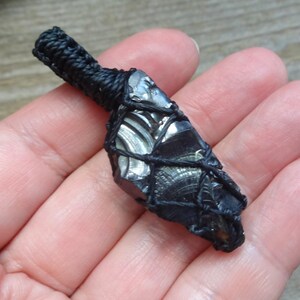 May include: A dark, reflective, irregular obsidian stone pendant encased in a black macrame cord netting. The stone features a smooth, glassy texture with subtle gray banding, secured by a thick, braided black cord loop at the top for hanging.