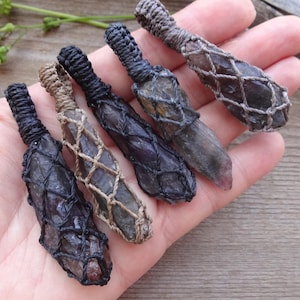 May include: Five raw crystal pendants encased in macrame netting. The stones are dark, translucent, and elongated. The netting is crafted from cord in shades of black, tan, and grey, with each pendant featuring a loop at the top for hanging.
