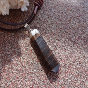 Puede incluir: Un colgante de piedra marrón y negra con un engaste plateado, colgado de un cordón de cuero marrón.