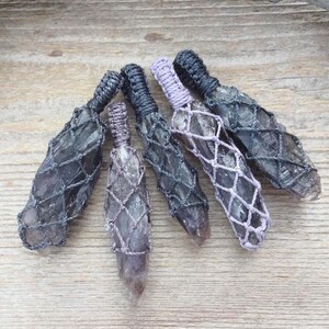 May include: Six dark gray crystal pendants wrapped in a woven net. The crystals are arranged on a light brown wooden surface, with a piece of driftwood in the background. The pendants have a variety of gray and purple tones.