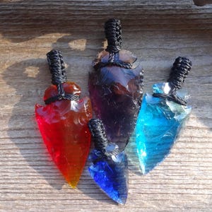 May include: Four colorful glass arrowhead pendants. The pendants are in shades of red, blue, and purple, with black woven cords at the top. The arrowheads are displayed on a wooden surface.