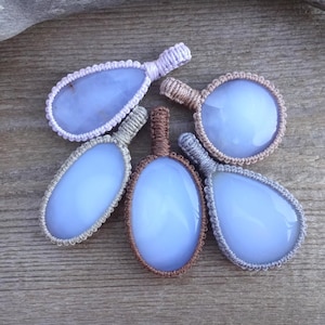 Peut inclure: Cinq pendentifs en calc&eacute;doine bleue avec des d&eacute;tails en macram&eacute; de diff&eacute;rentes formes : ovale, goutte d'eau et rond. Les pendentifs sont pos&eacute;s sur un fond en bois rustique, avec un morceau de bois flott&eacute; dans le coin sup&eacute;rieur gauche.