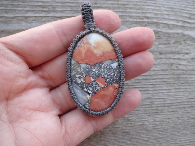 May include: A gray cord wrapped around a red, orange, and gray stone pendant. The stone has a marbled pattern.