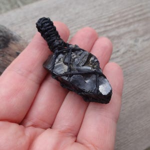 May include: A black crystal pendant wrapped in black cord. The crystal has a rough, textured surface with shiny, reflective areas. The cord is tightly woven at the top, forming a loop for hanging. The pendant is held in a hand.