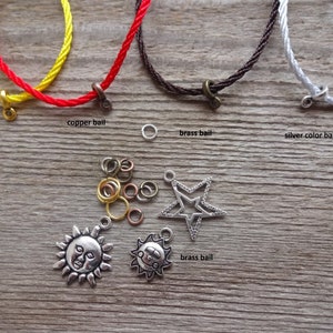 May include: A close-up of various cord colors and metal bails for jewelry making. The cords are red, yellow, brown, and white. The bails are gold, copper, and silver. There are also several jump rings and two charms: a sun and a star.