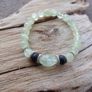 May include: A green gemstone bracelet with black beads and silver accents. The bracelet is made of round beads and has a larger, oval-shaped bead in the center.