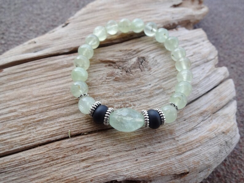May include: A green gemstone bracelet with black beads and silver accents. The bracelet is made of round beads and has a larger, oval-shaped bead in the center.