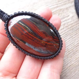 May include: Oval-shaped pendant with a polished, striped stone in shades of red, brown, and gold, encased in a black macrame setting. The pendant is held in a hand, showcasing the intricate detail of the stone and the macrame.