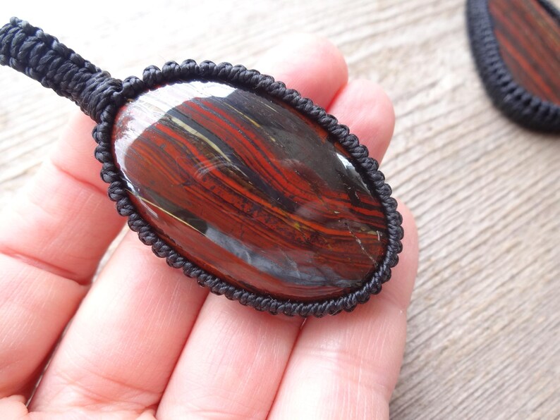 May include: Oval-shaped pendant with a polished, striped stone in shades of red, brown, and gold, encased in a black macrame setting. The pendant is held in a hand, showcasing the intricate detail of the stone and the macrame.