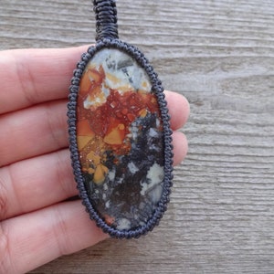 May include: A close-up of an oval-shaped pendant with a colorful, abstract pattern of red, orange, yellow, white, and black. The pendant is made of a natural stone and is strung on a dark gray cord.