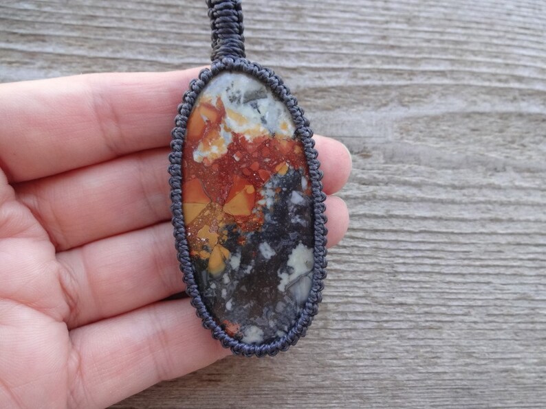 May include: A close-up of an oval-shaped pendant with a colorful, abstract pattern of red, orange, yellow, white, and black. The pendant is made of a natural stone and is strung on a dark gray cord.