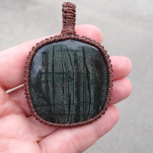 May include: A dark green stone pendant with a brown cord wrapped around the edge. The stone has a striated pattern.