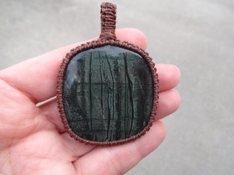 May include: A dark green stone pendant with a brown cord wrapped around the edge. The stone has a striated pattern.