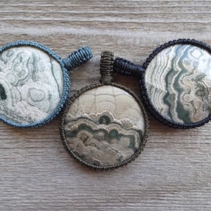 May include: Three circular pendants with distinctive stone patterns. Each pendant is framed by a woven cord and has a loop for suspension. The stones display shades of grey, green, and white, with the cord frames in blue, black, and olive green.