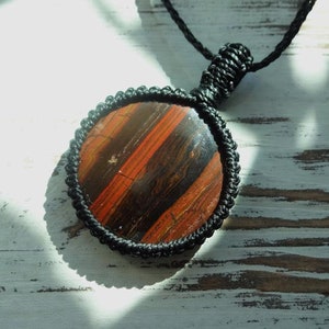 May include: Round pendant necklace featuring a polished stone with red and brown stripes, encased in a black macrame setting. The pendant hangs from a black cord, creating a striking contrast. The natural stone has a unique pattern.