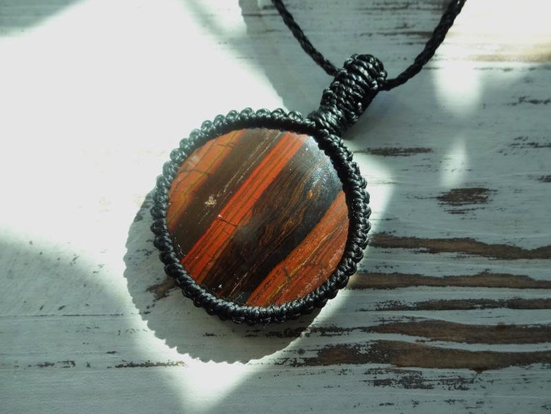 May include: Round pendant necklace featuring a polished stone with red and brown stripes, encased in a black macrame setting. The pendant hangs from a black cord, creating a striking contrast. The natural stone has a unique pattern.