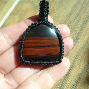 May include: A handcrafted pendant featuring a polished, rectangular stone with bands of red and brown hues. The stone is encased in a black woven macrame setting, with a loop for hanging. The pendant is held in a hand.