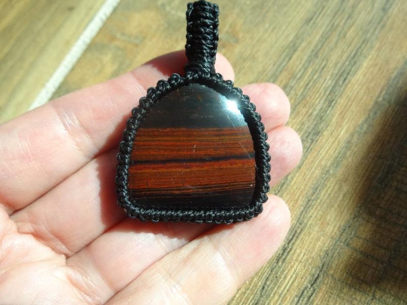 May include: A handcrafted pendant featuring a polished, rectangular stone with bands of red and brown hues. The stone is encased in a black woven macrame setting, with a loop for hanging. The pendant is held in a hand.