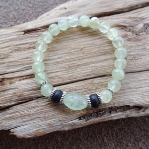 May include: A green gemstone bracelet with silver accents and black beads. The bracelet is made of round beads and a larger oval-shaped bead in the center.