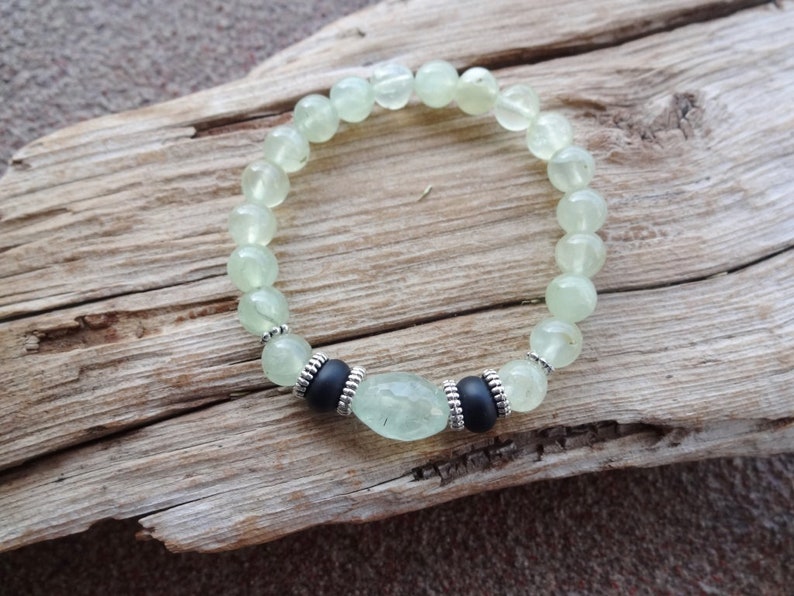 May include: A green gemstone bracelet with silver accents and black beads. The bracelet is made of round beads and a larger oval-shaped bead in the center.