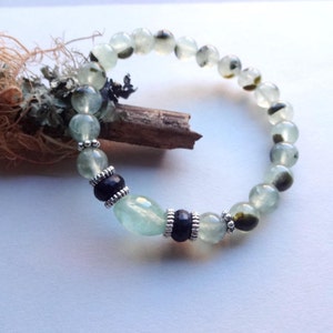 May include: A green and black gemstone bracelet with silver accents. The bracelet is made with round beads and two larger oval beads.