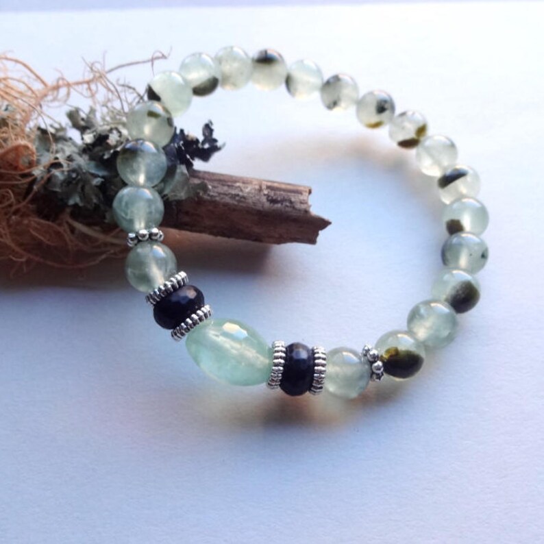 May include: A green and black gemstone bracelet with silver accents. The bracelet is made with round beads and two larger oval beads.