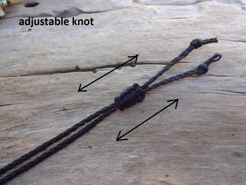 May include: A black cord necklace with an adjustable knot. The knot is tied in a way that allows the necklace to be adjusted to fit different neck sizes. The text "adjustable knot" is written on the wooden surface.