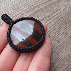 May include: A round pendant with a polished, striped gemstone in shades of red, brown, and white, set within a black woven macrame frame. The pendant has a small loop for hanging.
