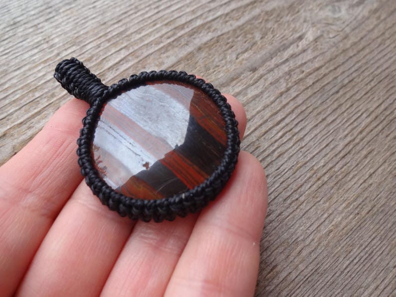 May include: A round pendant with a polished, striped gemstone in shades of red, brown, and white, set within a black woven macrame frame. The pendant has a small loop for hanging.