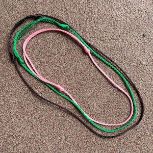 May include: Three cords of string in brown, green, and pink are tied together in a circle.