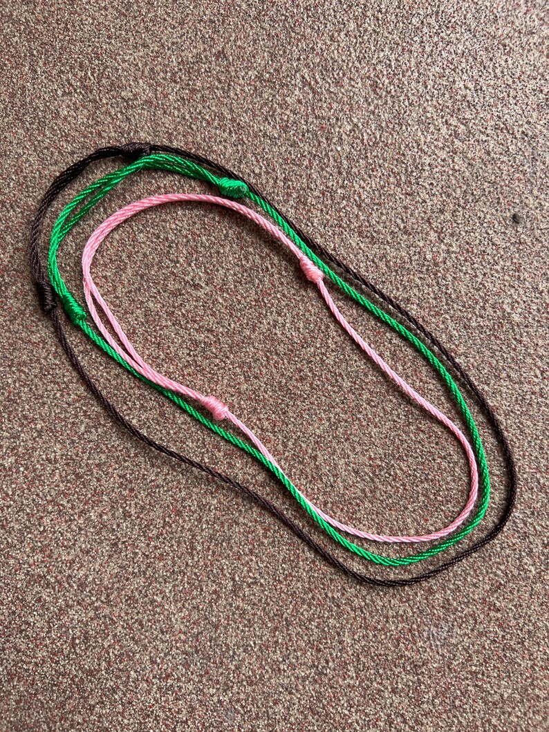 May include: Three cords of string in brown, green, and pink are tied together in a circle.
