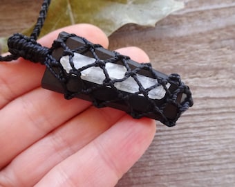 May include: A black rectangular pendant necklace with a white crystal center, encased in a black woven net. The necklace is suspended from a black cord. The pendant is held in a person's hand, showcasing the natural stone jewelry.