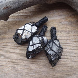 May include: Three pyrite pendants with a black woven cord. The pyrite stones are encased in a black net-like weave. The pendants are displayed on a wooden surface with a piece of driftwood in the background.