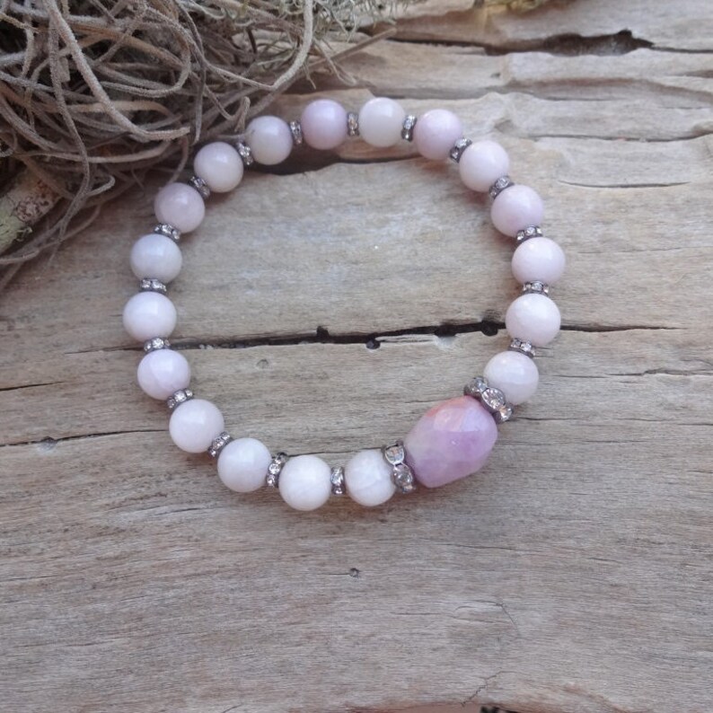 May include: A white and lavender beaded bracelet with a larger lavender bead in the center. The bracelet is made of natural stones and has a delicate, feminine look.