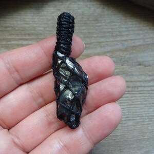 May include: A dark, metallic-looking raw stone pendant encased in a black macrame cord netting. The stone features a reflective, faceted surface with grey and black tones. The cord extends upward into a tightly woven loop for hanging.