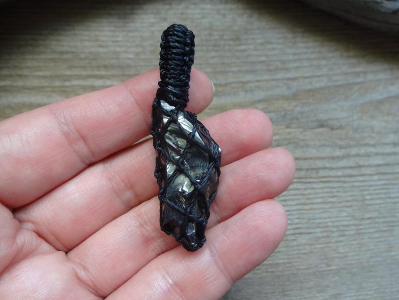 May include: A dark, metallic-looking raw stone pendant encased in a black macrame cord netting. The stone features a reflective, faceted surface with grey and black tones. The cord extends upward into a tightly woven loop for hanging.