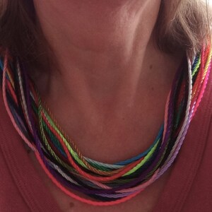 May include: A colorful multi-strand necklace made of thin cords in various shades of pink, purple, blue, green, yellow, and orange. The necklace is worn by a person with a pink shirt.