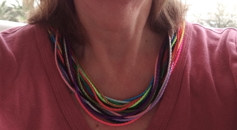 May include: A colorful multi-strand necklace made of thin cords in various shades of pink, purple, blue, green, yellow, and orange. The necklace is worn by a person with a pink shirt.