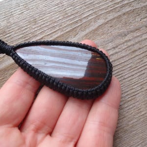 May include: A teardrop-shaped pendant with a polished, striped stone in shades of brown and white, encased in a black woven macrame setting. The pendant is held against a wooden background.