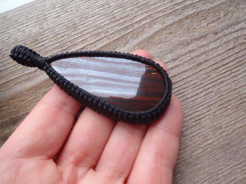 May include: A teardrop-shaped pendant with a polished, striped stone in shades of brown and white, encased in a black woven macrame setting. The pendant is held against a wooden background.