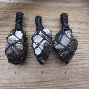 May include: Three raw, dark gray stones encased in black woven netting, each with a looped cord at the top. The stones have a rough, natural texture and are displayed on a wooden surface. Numbers 1, 2, and 3 are visible.