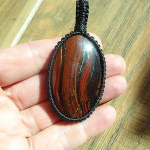 May include: Oval pendant featuring a polished stone with red and black striations, encased in a black woven macrame setting. The pendant has a loop for a necklace. The stone's pattern resembles wood grain.
