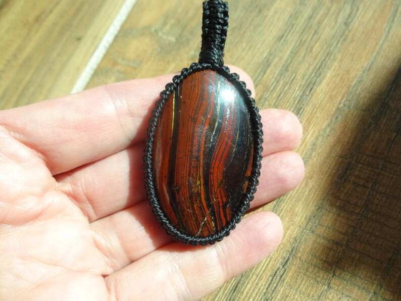 May include: Oval pendant featuring a polished stone with red and black striations, encased in a black woven macrame setting. The pendant has a loop for a necklace. The stone's pattern resembles wood grain.