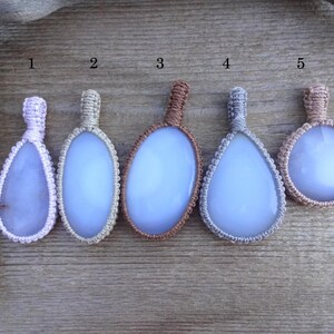 Peut inclure: Cinq pendentifs artisanaux avec des pierres bleu clair, chacun de forme unique et envelopp&eacute; de macram&eacute;. Les couleurs du macram&eacute; varient du violet clair au marron, sur un fond en bois. Ces pendentifs sont parfaits pour la cr&eacute;ation de bijoux.