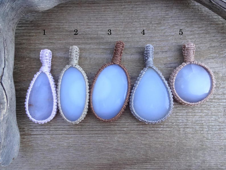 Peut inclure: Cinq pendentifs artisanaux avec des pierres bleu clair, chacun de forme unique et envelopp&eacute; de macram&eacute;. Les couleurs du macram&eacute; varient du violet clair au marron, sur un fond en bois. Ces pendentifs sont parfaits pour la cr&eacute;ation de bijoux.